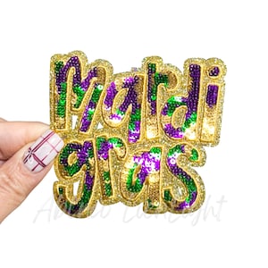 May include: A gold glitter "Mardi Gras" decoration with green, purple, and gold sequins. The letters are outlined in gold glitter. The decoration is held by a hand with a white and red nail design.