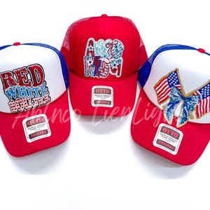 May include: Three red, white, and blue trucker hats with different patriotic designs. The first hat has the words "Red White Blue" in red, white, and blue letters. The second hat has the word "America" in red, white, and blue letters. The third hat has two American flags with a blue bow in the center.
