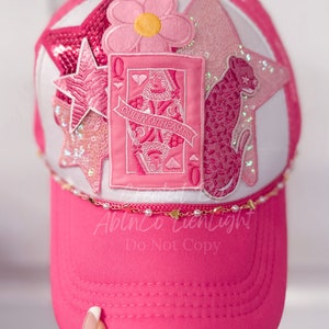 Puede incluir: Gorra de b&eacute;isbol rosa con una corona blanca y una visera rosa. La corona presenta una variedad de parches rosas y blancos, incluyendo una flor, una estrella, un leopardo y una carta de juego con la Reina de Corazones.