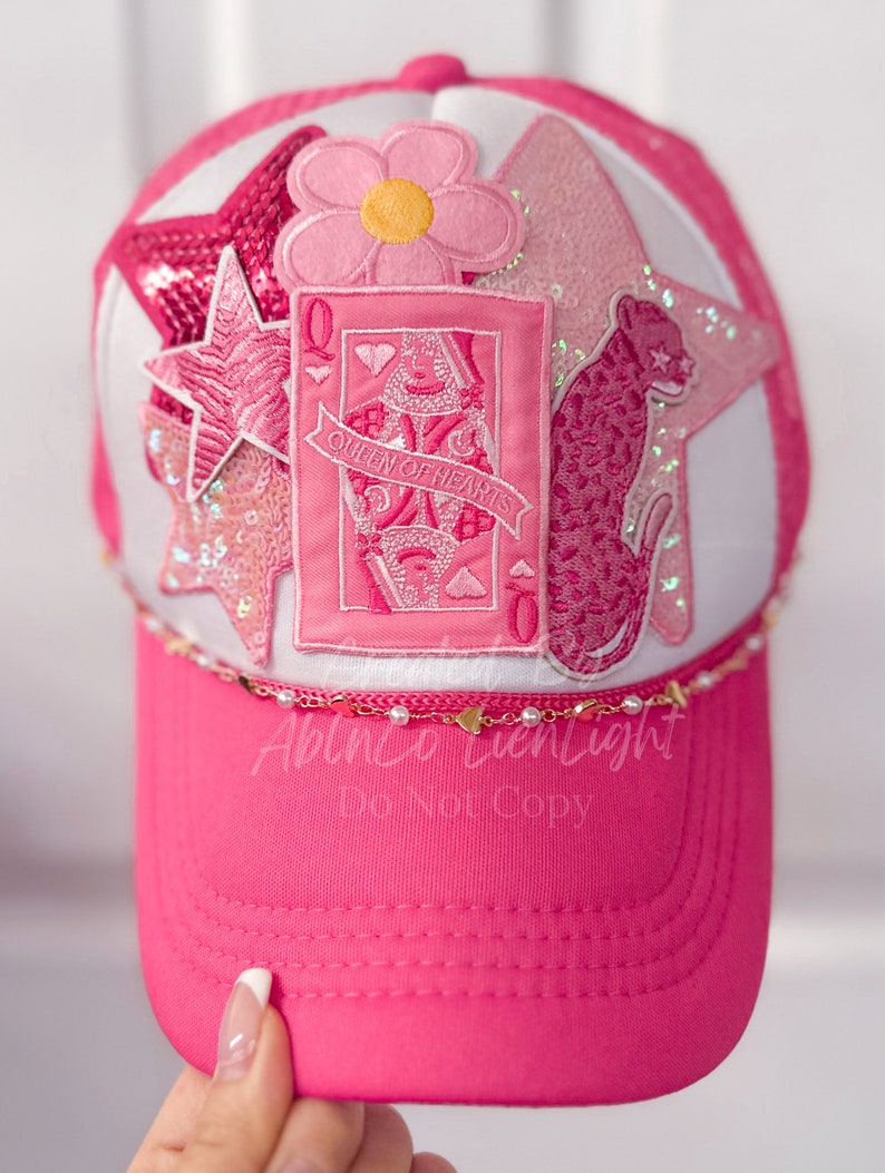 Puede incluir: Gorra de b&eacute;isbol rosa con una corona blanca y una visera rosa. La corona presenta una variedad de parches rosas y blancos, incluyendo una flor, una estrella, un leopardo y una carta de juego con la Reina de Corazones.
