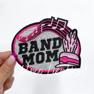 May include: A silver glitter iron-on patch with pink and black embroidery. The patch features the text "BAND MOM" with musical notes and a pink and black trumpet.