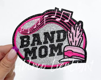 May include: A silver glitter iron-on patch with pink and black embroidery. The patch features the text "BAND MOM" with musical notes and a pink and black trumpet.