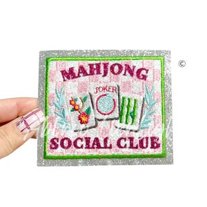Puede incluir: Parche bordado con las palabras "MAHJONG SOCIAL CLUB" en rosa, con un fondo de tablero de ajedrez. El parche tiene un borde plateado brillante y presenta diseños florales y de hojas. También se incluye la palabra "JOKER".
