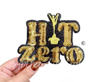 May include: A black and gold sequined patch with the words "HIT ZERO" and a gold figure. The letters are gold sequins on a black background. The figure is also gold. The patch is held by a hand.