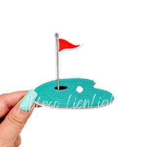 May include: A green and white embroidered patch of a golf hole with a red flag and a white golf ball.