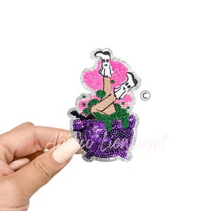 May include: A glittery patch featuring a witch's cauldron with a witch's legs and feet sticking out. The cauldron is purple with the words "so lien light" in a cursive font. The background is pink and green.