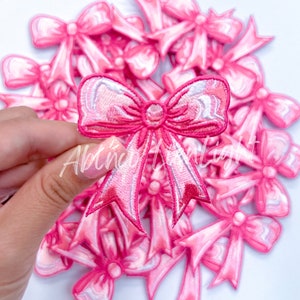 May include: A pile of pink satin bows with a white outline. The bows are arranged in a circular pattern with one bow in the center being held by a hand.