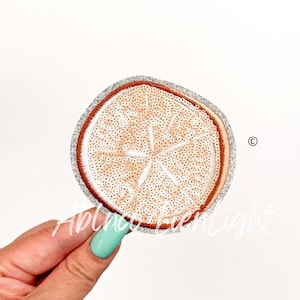 May include: A round, embroidered patch with a white, sequined orange slice design. The patch has a silver glitter border and a brown outline.