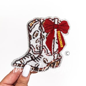 May include: A sequined patch shaped like a cowboy boot featuring a white background with brown football patterns. A large red bow is attached to the side of the boot, with the word TOUCHDOWN embroidered in yellow on one of the bow's tails.