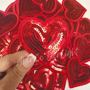 May include: A pile of red heart-shaped patches with sequins. Some of the hearts are plain red felt, while others are covered in red sequins.