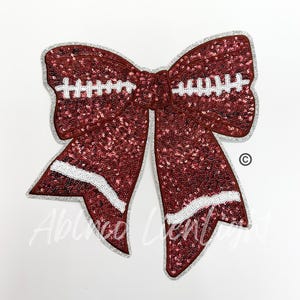 May include: A large red and white sequined bow with a football design. The bow is made of sequins and has a white outline. The football design is on the center of the bow and has white stitching.