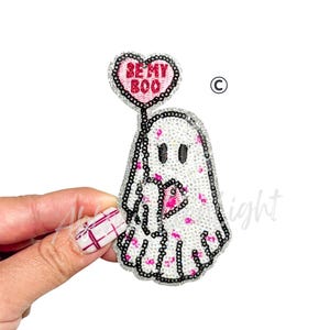 May include: A sequined ghost-shaped patch with a heart-shaped balloon that reads "BE MY BOO". The ghost is white with pink accents and black details. The patch is held up by a hand against a white background.