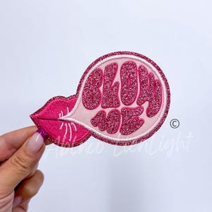 May include: Pink iron-on patch with a pink glitter speech bubble that says "Blow Me" and a pink glitter lip blowing into the bubble.