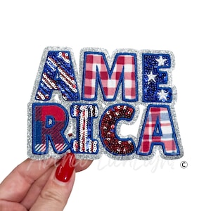 May include: A patriotic patch spelling "AMERICA" in red, white, and blue. The letters are decorated with sequins, plaid patterns, and star designs, outlined with silver glitter. The patch is held up by a hand with red nail polish.