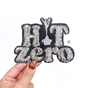 May include: A black sequined patch with the words "HIT ZERO" in silver sequins. A silver cheerleader figure is in the center. The patch has a glittery border and is held by a hand.