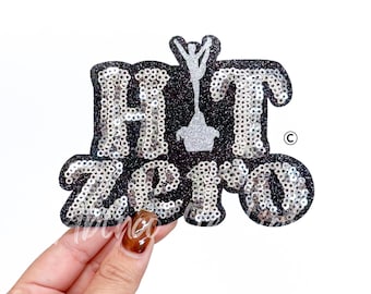 May include: A black sequined patch with the words "HIT ZERO" in silver sequins. A silver cheerleader figure is in the center. The patch has a glittery border and is held by a hand.