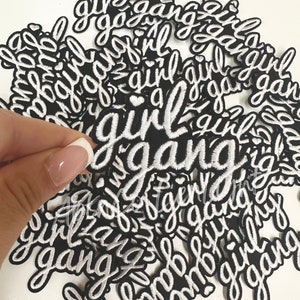 May include: A pile of black iron-on patches with white embroidery that reads "girl gang".