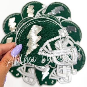 May include: A stack of green and white felt patches shaped like football helmets with a silver sequin lightning bolt on each patch.