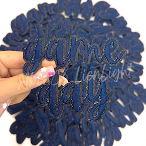 May include: A close-up of a hand holding a navy blue glitter iron-on patch that says "game day".