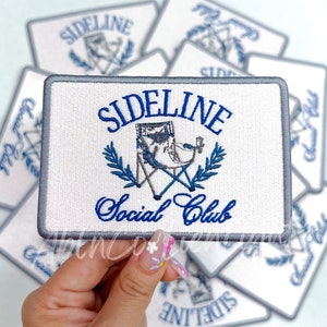 Puede incluir: Un parche blanco bordado con un borde gris. El parche presenta una ilustración azul y blanca de una silla plegable con una corona azul a cada lado. El texto "Sideline Social Club" está bordado en azul debajo de la silla.