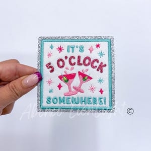 May include: Embroidered iron-on patch with a silver glitter border. It features two pink martini glasses with green olives and the text "It's 5 o'clock Somewhere!"