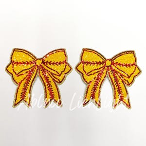 May include: Two yellow sequined bow appliques with red accents and gold trim. The bows are symmetrical and have a classic bow shape. The appliques are on a white background.