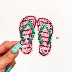 May include: Two pink sequined flip-flops with green straps. The flip-flops have a light pink border and are on a white background. The flip-flops are held by a hand with a turquoise nail polish.