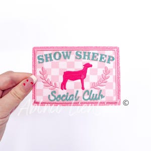 May include: Pink and white checkered patch with a pink silhouette of a sheep and the text "Show Sheep Social Club" in teal.