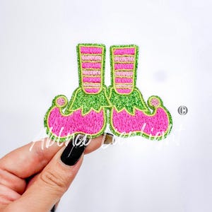May include: A pair of pink and green elf shoes with white stripes. The shoes are made of felt and have a glitter finish.