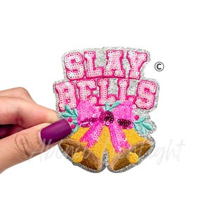 May include: A decorative patch with the words "SLAY BELLS" in pink sequins. The patch features two yellow bells with brown accents, a pink bow, and green holly leaves. The patch is held by a hand.