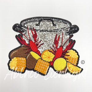 May include: A silver pot with red crawfish, corn on the cob, potatoes, and lemons. The pot is covered in sequins.