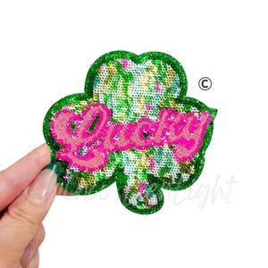 May include: A sequined shamrock patch with the word "Lucky" in pink script. The shamrock has a green glitter border and is filled with green, gold, and white sequins. The patch is held by a hand.
