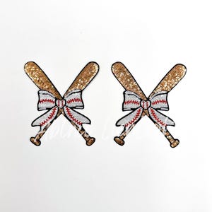 May include: Two gold sequin baseball bats crossed with a white bow with red stripes in the center. The bats are decorated with white and red baseball stitching.