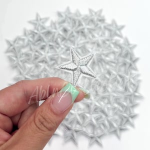 May include: A pile of silver glitter star iron-on patches. The patches are about 1 inch in size and are perfect for adding a touch of sparkle to any project.