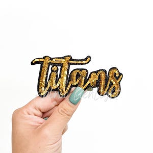 May include: A gold sequined patch with the word "Titans" in a cursive font. The patch has a black border and is held against a white background. The sequins give the patch a shiny, textured appearance.