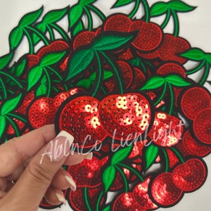 May include: A pile of red and green cherry-shaped patches with sequins. The cherries are embroidered with black thread and have green stems and leaves.