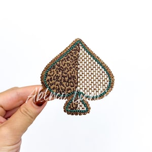 May include: A decorative spade-shaped patch with a leopard print pattern on the left side and a woven design on the right. The patch is outlined in gold and green, held against a white background.