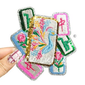 May include: Embroidered patch featuring a peacock with a colorful tail, surrounded by other embroidered elements. The patch has a silver glitter border and is held by a hand. The peacock is blue, and the surrounding elements are pink, green, and yellow.