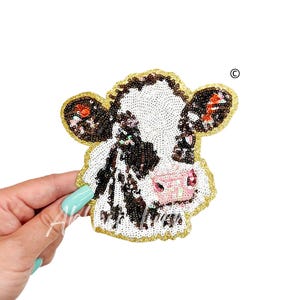 May include: A black and white cow face patch made of sequins with a pink nose and gold glitter around the edges.