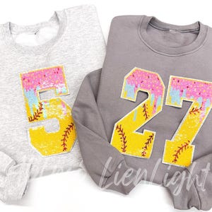 May include: Two sweatshirts, one light gray and one dark gray, each with a large, colorful number patch. The patches feature a yellow baseball design with pink, blue, and white accents. The light gray sweatshirt has the number 5, and the dark gray has the number 27.