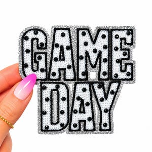 May include: A decorative patch featuring the text GAME DAY in block letters. The letters are white with a silver glitter border and are embellished with scattered black circular studs. The entire design is outlined in a thick black stitched border.
