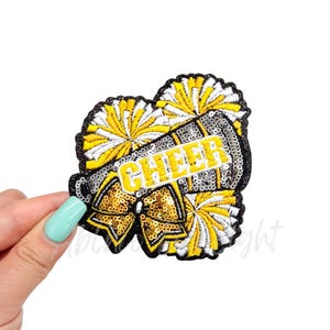May include: A yellow and white cheerleading pom pom with a black and silver megaphone with the word "CHEER" on it. The megaphone has a gold bow on it.