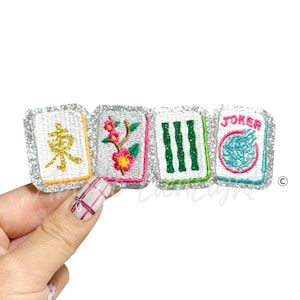 May include: Four embroidered Mahjong tile patches with silver glitter borders. The tiles feature various designs, including Chinese characters, flowers, and the word "JOKER". The patches are in shades of white, green, pink, and yellow.
