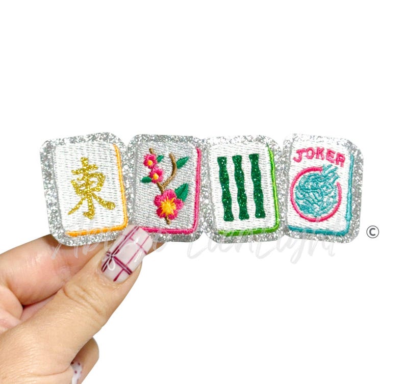 K&ouml;nnte beinhalten: Vier gestickte Mahjong-Kachel-Patches mit silbernen Glitzerborten. Die Kacheln weisen verschiedene Designs auf, darunter chinesische Schriftzeichen, Blumen und das Wort "JOKER". Die Patches sind in Wei&szlig;-, Gr&uuml;n-, Rosa- und Gelbt&ouml;nen gehalten.