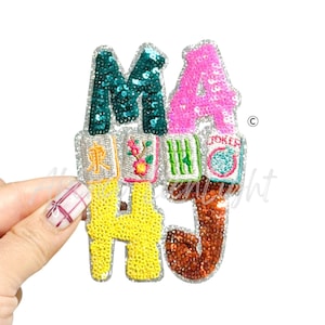 May include: A sequined patch spelling out "MAHJ" in teal, pink, yellow, and brown. The letters are embellished with sequins and embroidered details. The patch includes images of Mahjong tiles.