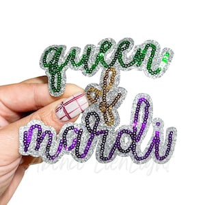 May include: A glittery, sequined accessory with the words "queen of mardi" in a cursive font. The word "queen" is green, "of" is gold, and "mardi" is purple. The letters are outlined in silver.
