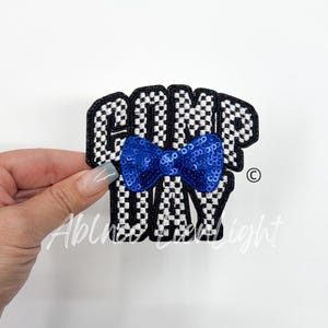 May include: A black and white checkered patch with the words "GOMP DAY" in a bold font. A blue sequined bow is centered on the patch. The patch is held by a hand.