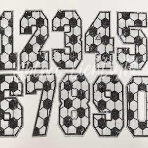 May include: Ten iron-on patches in the shape of numbers 1 through 10. Each number is made of a black and white soccer ball pattern.