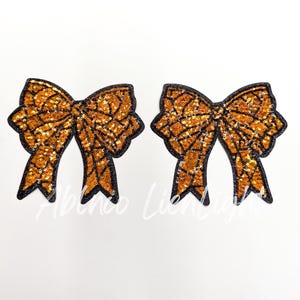 May include: Two orange sequined bow appliques with black outlines and a spiderweb design. The bows are on a white background. The text "Albenco Lien Light" is visible at the bottom.