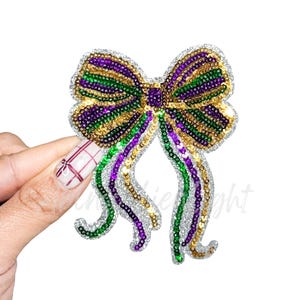 May include: A sequined bow in purple, green, and gold, with silver trim and dangling ribbons. The bow is held against a white background. The design is reminiscent of Mardi Gras.